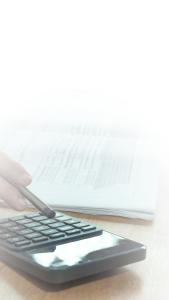 A hand holding a pen presses buttons on a calculator, with an open document containing charts and graphs blurred in the background. The image has a light, faded effect at the top.