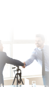 Two people in business attire shaking hands in an office with large windows, a tripod and a glass of water visible in the background. The image is brightly lit with a faded effect at the top.