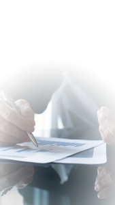 A person's hands holding a pen hovering over paper displaying a bar chart, suggesting business analysis or financial review. The top of the image is faded white.