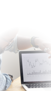 A person points at a laptop screen displaying a line graph and bar chart with monthly data, suggesting business analysis or financial review. The top of the image is faded white.