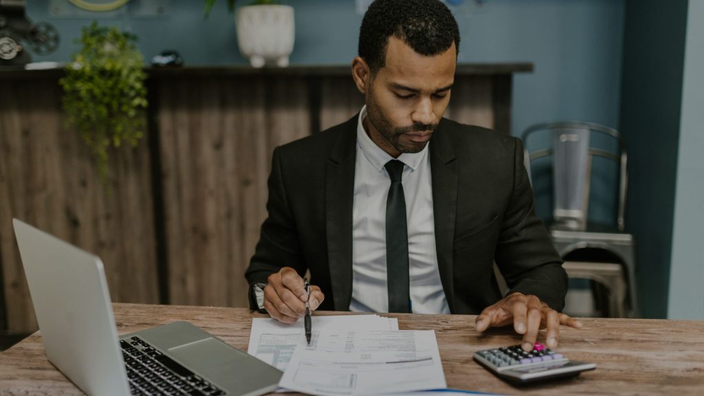 Male accountant in a black suit and tie behind a desk looking at paperwork and typing into a calculator