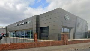 Land Rover dealership building with large glass windows, green Land Rover branding, and a low red-brick wall along the foreground under a partly cloudy sky.